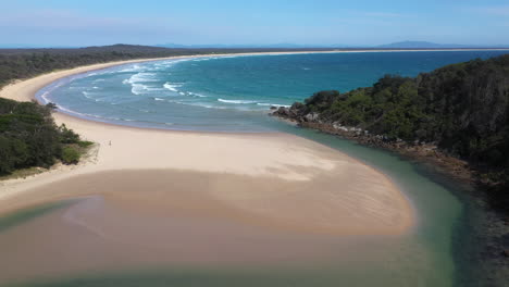 Wide-descending-shot-of-the-South-Pacific-Ocean-and-Korogoro-Creek-with-wind-blowing-sand-across-a-sand-bar-at-Hat-Head-New-South-Wales,-Australia