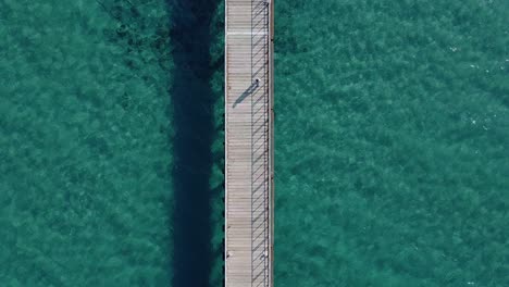 Overhead-drone-view-of-a-person-walking-along-a-long-wood-pier-surrounded-by-sparkling-tropical-waters