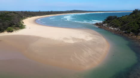 Cinematic-tilting-up-drone-shot-of-the-South-Pacific-Ocean-and-Korogoro-Creek-with-wind-blowing-sand-across-a-sand-bar-at-Hat-Head-New-South-Wales,-Australia
