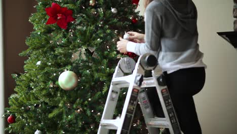 Girl-On-Step-Ladder-Decorating-Christmas-Tree-With-Christmas-Lights,-Balls,-And-Poinsettia-Flower-For-Christmas-Season