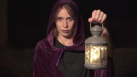 Mysterious-role-play-woman-wearing-a-purple-cape-and-hood-and-holding-a-lantern-at-night