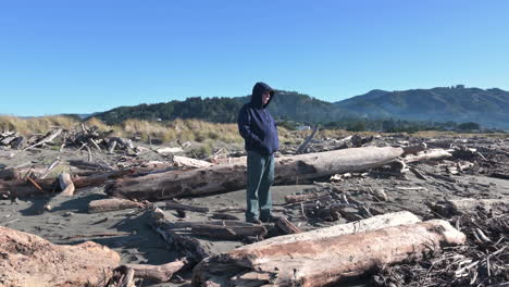 Old-Man-Wearing-Blue-Hoody-Sweater-Standing-At-Riverbank-With-Deadwoods-During-Summer-In-Gold-Beach,-Oregon