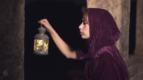 Young-mysterious-woman-wearing-a-cloak-and-hood-holding-a-lantern-looking-for-something-in-the-dark-at-night