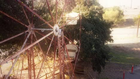 Aerial-around-a-rusting-ferris-wheel-pulling-back-to-reveal-an-abandoned-parking-lot-and-the-top-of-a-tall-palm-tree