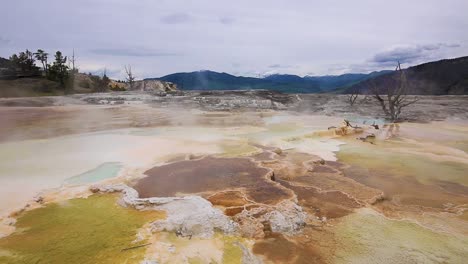 Geothermal-hot-springs-with-steam-cloud-rising-and-mountains-in-the-background-at-Yellowstone-National-Park,-Wyoming,-USA