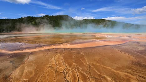 Static-view-of-geothermal-geyser-and-hot-spring-with-blue-steam-cloud-rising-and-mountains-in-the-background-at-Yellowstone-National-Park,-Wyoming,-USA