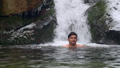 Handsome-man-ice-swimming-in-subzero-cold-river-by-the-beautiful-waterfall-in-snowy-mountains-during-winter-day---static-shot,-slow-motion