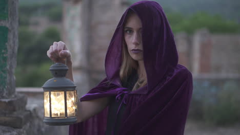 Mysterious-young-woman-wearing-a-cloak-and-a-hood-covering-her-head-staring-at-the-camera-and-holding-an-old-lantern