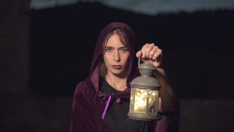 Mysterious-young-woman-in-a-purple-cloak-and-a-hood-staring-at-the-camera-and-using-an-old-lantern-at-night