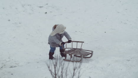 Young-girl-slowly-pushes-wooden-sled-up-a-snow-covered-hill,-Wide-shot