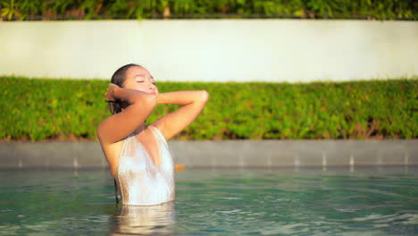 Slow-motion-shot-of-a-attractive-young-woman-standing-half-submerged-in-the-pool-touching-her-wet-long-hair-with-her-eyes-closed-and-then-slowly-opening-them-with-delight-and-smile-on-her-face