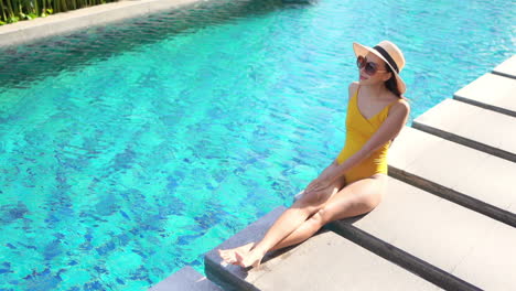 Pretty-young-woman-sitting-near-a-swimming-pool,-wearing-one-piece-yellow-swimsuit,-sun-hat-and-sunglasses,-sunbathing-and-enjoying-her-vacation