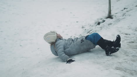 Girl-rolls-down-snowy-hill-after-struggling-to-slide-on-back,-Slow-Motion