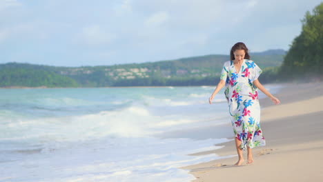 Beautiful-young-Asian-model-walking-barefoot-on-the-white-sand-island-beach-wearing-a-sundress-when-foamy-waves-crashing,-slow-motion-daytime