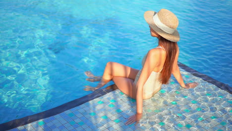 Attractive-Female-in-Swimsuit-and-Hat-Sitting-in-Blue-Pool-on-Sunny-Day