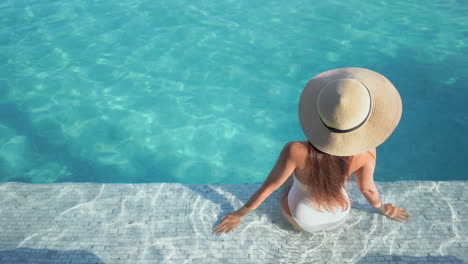 Glamour-Female-in-Swimsuit-and-Summer-Hat-Sitting-on-Swimming-Pool-Edge-on-Sunny-Day