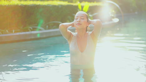 Sexy-Petite-Asian-Female-in-Swimming-Pool-With-Hands-in-Wet-Hair-Looks-At-Camera-Slow-Motion-on-Bright-Summer-Day