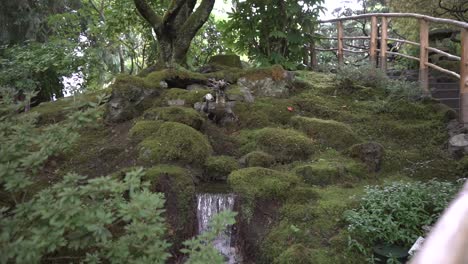 Tiro-De-Seguimiento-De-Rocas-Cubiertas-De-Musgo-Que-Rodean-Una-Cascada-En-Miniatura-En-Un-Jardín-Japonés