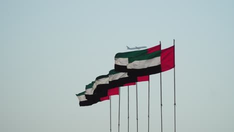 Banderas-De-Los-Emiratos-árabes-Unidos-Ondeando-En-El-Viento,-El-Símbolo-Nacional-De-Los-Emiratos-árabes-Unidos,-Aviones-Comerciales-De-Pasajeros-De-Los-Emiratos-De-Airlin-Con-Base-En-Dubai-Que-Pasan-En-El-Fondo,-4k-Vidoe