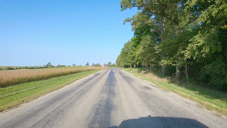 POV-driving-on-rural-county-road-with-no-center-line-past-maturing-fields,-farmyard,-and-bicyclist-in-rural-Iowa-on-a-sunny-early-autumn-day