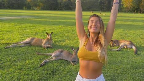 Beautiful-Girl-In-Bralette-Raised-Both-Hands-With-Eastern-Grey-Kangaroos-Resting-In-The-Background---Gold-Coast,-QLD,-Australia