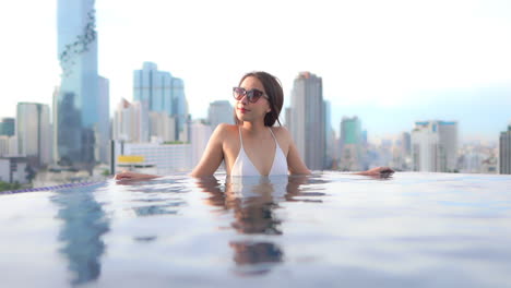 With-a-background-of-a-modern-urban-skyline,-a-pretty-young-woman-leans-against-the-edge-of-a-resort-rooftop-infinity-pool