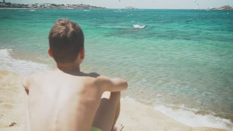 Young-boy-sitting-on-a-crystal-beach-in-a-Greek-Island