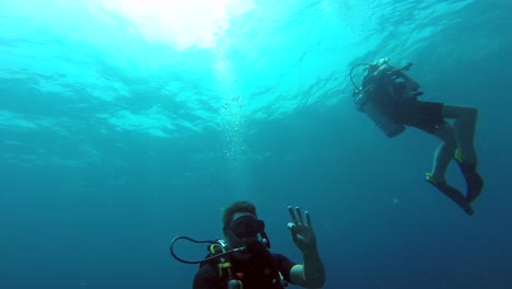Pair-of-scuba-divers-floating-just-beneath-the-ocean-surface,-with-one-signaling-to-a-co-diver