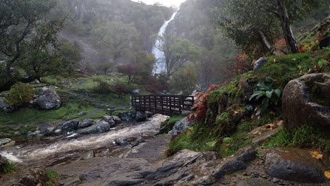 Waterfall-rocky-river-flowing-water-slow-motion-cascades-under-wooden-bridge-in-national-park-wide-shot