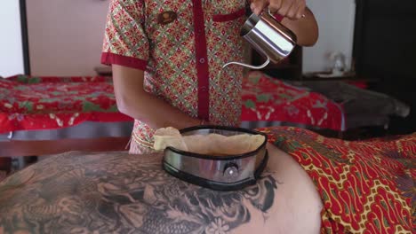 Spa-therapist-slowly-pours-oil-into-the-wax-lined-container-on-the-back-on-a-patient-enjoying-a-Kati-Vasti-Ayurvedic-treatment