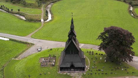 Aerial-View,-Hopperstad-Black-Wooden-Stave-Catholic-Church-and-Cemetery,-Norway,-Ancient-Religious-Norwegian-Monument-in-Green-Summer-Landscape,-Drone-Shot