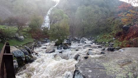 Cascadas-De-Agua-Que-Fluyen-Rápidamente-A-Cámara-Lenta-Bajo-Un-Puente-De-Madera-Desde-La-Cascada-Del-Valle-Del-Bosque