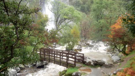 Idyllischer-Holzsteg-über-Dem-Wasserfall-Im-Herbsttal,-Der-In-Einen-Mächtigen-Fließenden-Fluss-Kaskadiert