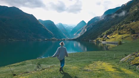 Slow-motion-shot-of-a-girl-running-and-walking-over-a-grass-field-towards-beautiful-scenic-view-of-a-large-lake-and-tall-mountains-in-Olden,-Norway