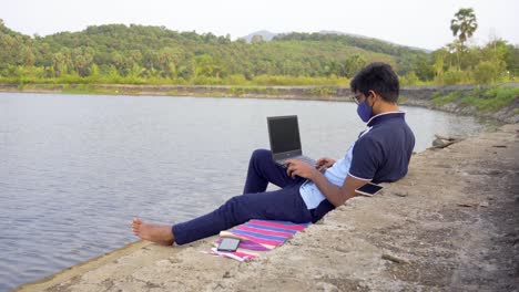 working-remotely-from-laptop-waring-mask-location-lakeside-India-new-normal-wide-shot-relaxed