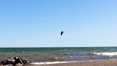 A-lone-kiteboarder-skillfully-traversing-a-rocky-shoreline,-on-a-beautify,-virtually-cloudless-day