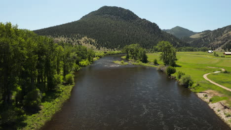 River-flowing-through-green-sunny-landscape-with-mountains,-drone