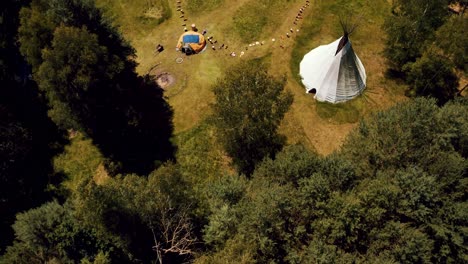 Drone-Flies-over-a-beautiful-Dense-Green-forest-to-reveal-a-large-outdoor-event-space-with-giant-Teepee,-native-american-community