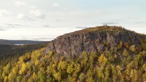 Aerial-orbit-pan-shot-of-rocky-mountain-peak-surrounded-by-pine-trees-and-lush-vegetation-in-the-Fall-and-revealing-vast-boreal-forest-disrupted-by-nordic-river-in-the-horizon,-in-Sweden,-Lapland