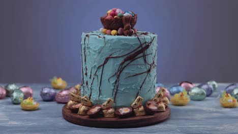 Trato-Tradicional-De-Pascua:-Delicioso-Pastel-De-Nido-De-Pascua-Azul-Decorado-Con-Conejos-Dorados-Y-Rodeado-De-Huevos-De-Pascua-De-Colores-Sobre-Una-Mesa-Azul