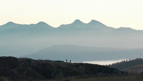 Shot-of-people-walking-over-a-small-mountain-in-the-distance,-large-mountains-in-the-background,-Norway,-Molde