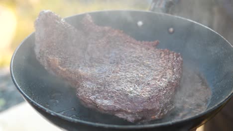Close-up-shot-of-Prime-rib-entrecôte-being-flipped-and-roasted-brown-in-sizzling-hot-pan,-with-the-aromas-condensing-and-disappearing-into-a-camping-outdoor-environment