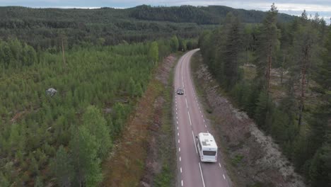 Aerial-tracking-shot-behind-camper-van-crossing-along-forest-road,-surrounded-by-high-pine-trees,-in-mountainous-environment