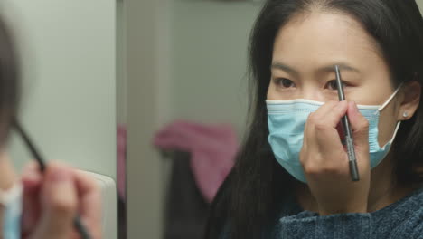 Young-Asian-American-woman-with-face-mask-applying-eyebrow-makeup
