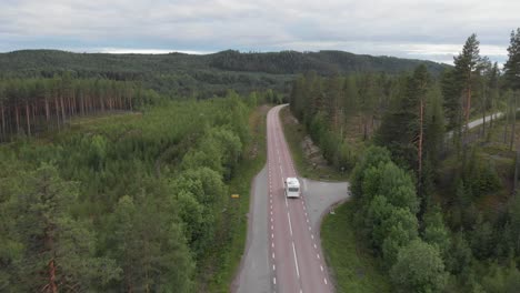 Aerial-tracking-shot-of-mountainous-lonely-road-splitting-green-nordic-forest,-while-chasing-white-camper-van-going-on-an-adventure