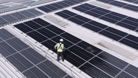 Male-Worker-Cleaning-Solar-Panel-On-The-Roof-Of-A-Large-Warehouse-Using-An-Electric-Rotary-Brush-Motor-With-Double-Heads---pullback-drone-shot