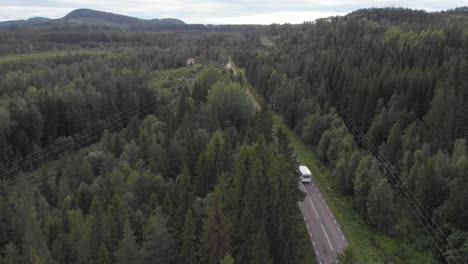 Aerial-wide-shot-of-white-camper-van-driving-down-road,-gently-rising-up-revealing-lush,-dense,-dark-green-nordic-forest-full-of-coniferous-trees