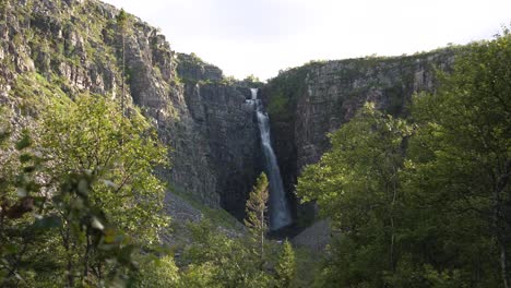 Wide-long-shot-of-Njupeskär-waterfall,-plunging-fresh-water-between-eroded-canyon,-at-Fulufjället-National-Park,-surrounded-by-spruce-trees-shrouded-in-sunlight,-in-Särna,-Sweden