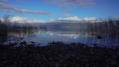 Dämmerung-Am-Felsigen-Ufer-Des-Ruhigen-Sees-Mit-Wasser,-Das-Weiße-Wolken-Am-Blauen-Himmel-Reflektiert