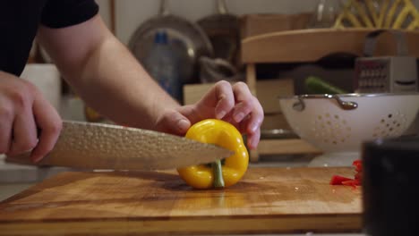 Chef-cuts-big-yellow-color-pepper-with-steel-knife-on-wooden-cut-board-on-a-kitchen-table